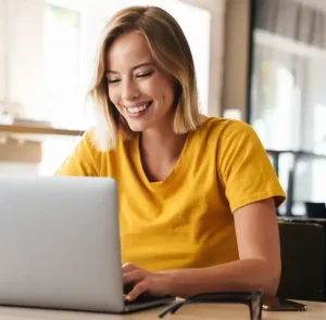 A smiling white woman with short blonde hair and a yellow tshirt works on a laptop