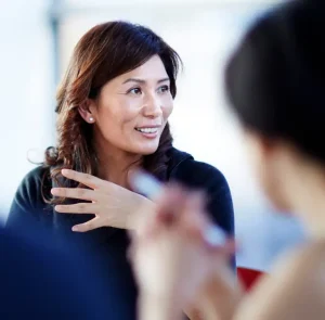 A woman with medium-length brown hair smiles and gestures with her hand while speaking to two blurred people in the foreground, suggesting a lively conversation or discussion.
